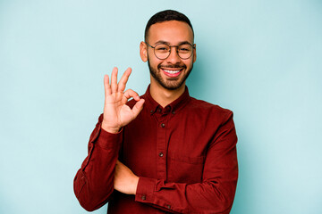 Wall Mural - Young hispanic man isolated on blue background winks an eye and holds an okay gesture with hand.