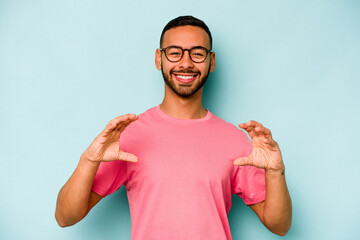 Wall Mural - Young hispanic man isolated on blue background holding something with palms, offering to camera.