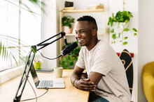 Happy young african man making audio podcast from home - Content creator recording radio show using professional microphone at small broadcast studio
