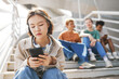© Seventyfour - Portrait of Asian teenage girl holding smartphone outdoors while sitting on metal stairs with group of friends in background, copy space