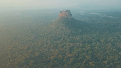 Wall Mural - Aerial drone shot of Sigiriya Lion Rock in Sri Lanka at sunrise with light mist and haze filled skies overlooking natural tropical forest and lush mountain ranges with orange blue tones