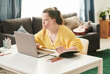 Portrait Of Young Female University Student With Down Syndrome Sitting At Table At Home Using Laptop To Do Homework