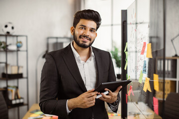 Portrait of indian executive manager wearing formal clothes, holding digital tablet and standing near glass wall with sticky papers. Concept of business, people and technology.