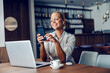 © Dusan Petkovic - A happy elegant businesswoman with earphones sits in a cafe and using her phone for a video call. She is taking a break from work. There is a laptop on the table. A businesswoman having a video call
