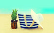 Summer vacation concept - bag with straw hat and pineapple on beach on blue sea background