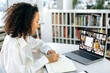 © Kateryna - Online learning concept. Positive smart dark-skinned girl, sits in the library, in front of the laptop screen, goes through online video training, listens attentively the lecture, smiles, takes notes