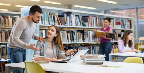 Portrait of two young adult students studying at library using books and laptop, working on common project