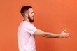 © khosrork - Side view portrait of positive friendly handsome bearded man holding out his hand to side, giving handshake, wearing pink T-shirt. Indoor studio shot isolated on orange background.