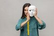 Portrait of strict bossy woman covering half of face with white mask, multiple personality disorder, wearing casual style jacket. Indoor studio shot isolated on gray background.