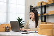 © Chanakon - Young asian entrepreneur woman checking product on stock take note with laptop. Business woman working SME online at home. Check address on parcel. Small business owner start up freelance concept.