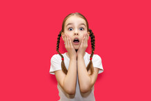 Girl Child With Pigtails On A Red Background With A Surprised Look Holds On To Her Face. Shocked Little Girl With Big Eyes And Open Mouth. Child With A Grimace Of Shock And Surprise