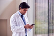 Male Doctor Wearing White Coat Standing In Hospital Corridor Looking At Mobile Phone