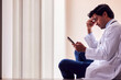 © Monkey Business - Stressed Male Doctor Wearing White Coat Sitting On Floor In Hospital Corridor With Mobile Phone