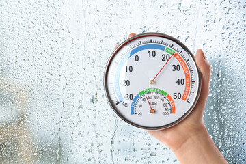 Woman holding hygrometer with thermometer near window on rainy day, closeup. Space for text