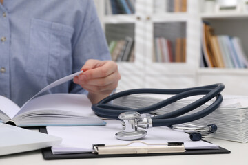 Woman with book, stethoscope and clipboard at white table indoors, closeup. Medical education