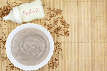 Wall Mural - Buckwheat porridge for the baby from ground cereals in a white bowl, a bottle of milk on a brown bamboo background. Space for the text. The first complementary food of a child, baby nutrition.