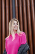 Happy beautiful blonde woman with a smile in a fashionable pink sweater and jacket walking on the street near the metal wall