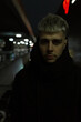 Fashion urban night portrait of a handsome serious brutal guy in a black jacket with hood stands on the street at night