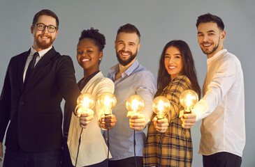 Wall Mural - Group portrait of happy intelligent young people with glowing lit lightbulbs. Smiling multiethnic business collaboration team with shining light bulbs looking at camera sharing creative idea with you