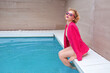 portrait of marvelous stylish redhead curly young woman sitting in fuchsia jacket, stylish sunglasses and black underpants by the swimming pool. Fashion, style, leisure, recreation, travel, make up co
