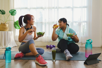 Fitness and Healthy food concept. Two Asian women body size is different in sportswear sitting smiling enjoys while eating healthy food together and happiness after fitness exercising at home.