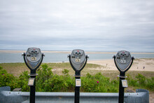 Three Tower Viewer Binoculars Mounted On A Stalk Looking Toward The Camera Lens Near A Sandy Beach Under A Cloudy Blue Sky In MA, USA