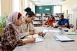 © pressmaster - Group of ethnically diverse students sitting at table watching educational videos on digital tablet and doing writing task