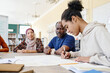 © pressmaster - Young Black immigrant sitting at table writing something on handmade educational English language poster during lesson