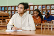 © pressmaster - Pensive young Black man sitting at desk in classroom having lesson at international school for immigrants listening to teacher and making notes