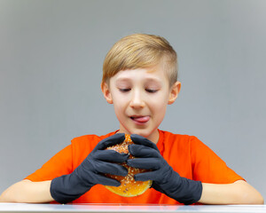 Wall Mural - a boy sitting at a table in black disposable gloves holds in front of him a huge burger sprinkled with sesame seeds a boy looking at a burger licks his lips