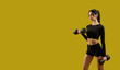 © serhiibobyk - Side view of pretty slim girl standing, raising dumbbells. Brunette young woman making step, working out, stretching, looking forward. Isolated on yellow studio background.