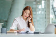 © amnaj - Happy secretary Asian business woman charming sitting smiling and talking on the smartphone in the office.