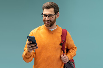 Portrait of cheerful brunette student man with beard using cellphone and smiling, reading good news message, enjoying mobile application. Indoor studio shot isolated on blue background