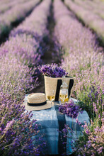 Two Glasses With White Wine And Bottle On A Background Of A Lavender Field. Straw Hat And Basket With Flowers Lavender On A Blanket On Picnic Table. Romantic Evening In Sunset Rays. Time For Dinner.