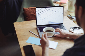 Man holding coffee in cafe, working with drawing on laptop. Freelancer in cafe. View over shoulder.