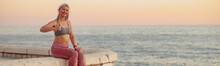 Sporty Woman Sitting On Ledge Of Waterfront On Background With Horizon Of Sea With Pink Sky, Smiling, Showing Thumbs Up