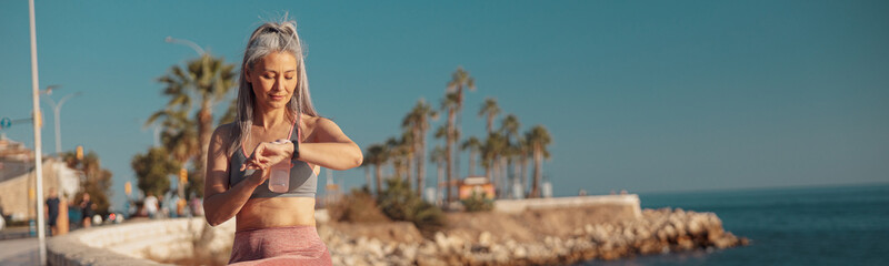strong lady in sports clothes sitting by the seashore, holding bottle of water and looking at the wa