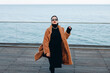 beautiful fashion model girl in black glasses and beige coat posing on the terrace of a restaurant overlooking the sea