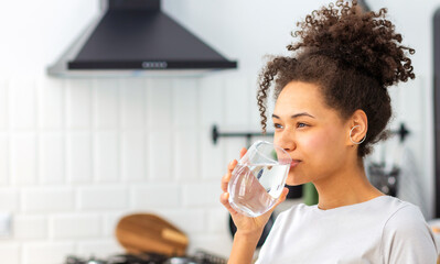 Healthy lifestyle concept. Beautiful young Afro American woman drinking clean water standing at home in the kitchen, copy space