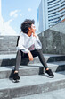 © anderson - Afro teenage female person sitting on stairs of building in city wearing sportswear with wind in her hair on sunny day