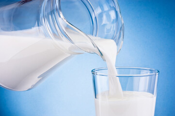 Wall Mural - Milk poured from jug into glass on blue background