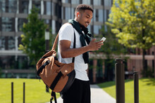Young black man in earbuds using phone walking in city