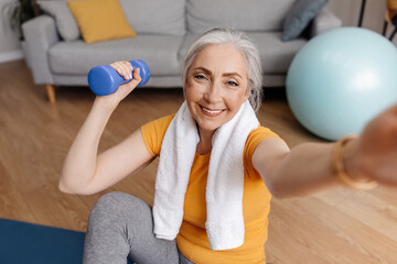 Canvas Print - Excited senior woman taking selfie while exercising with dumbbells at home, sitting on yoga mat