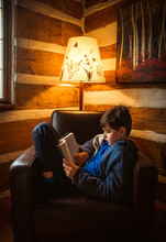 Boy Reading Book In Chair In The Corner Of A Rustic Log Cabin.