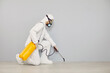 Pest control guy cleaning home from insects. Man in white protective suit crouching near wall and spraying floor with cockroach insecticide from yellow bottle for safe living environment inside house