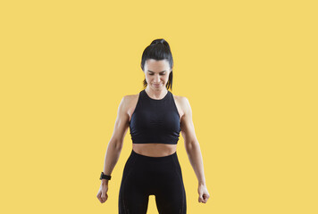 Wall Mural - Strong muscular sportive woman in sportswear isolated on yellow studio background feel powerful and motivated. Fit toned female athlete or coach show muscles. Sport and physical activity.