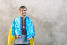 Young Smiling Ukrainian Dresses The Blue And Yellow State Flag Of Ukraine On His Shoulders. Man In Traditional Embroidered Shirt On Background Of Textured Concrete Wall. Constitution Day. Copy Space.