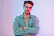 Men's studio portrait of a handsome hipster guy with a haircut and vintage glasses in a casual denim outfit with a jeans shirt in the studio with pink and blue light