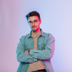 Creative portrait of fashionable handsome young artist man with vintage eyewear in denim outfit with jeans shirt stands in studio with pink and blue lights