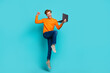 Full length photo of lucky pretty guy dressed orange sweater jumping typing modern gadget isolated turquoise color background